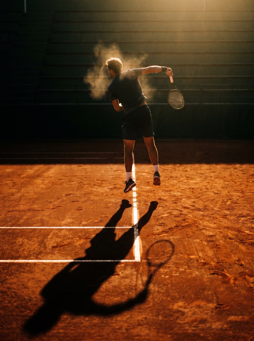 A clay court at golden hour — a single player mid-serve, long shadow, crisp line markings and clean light